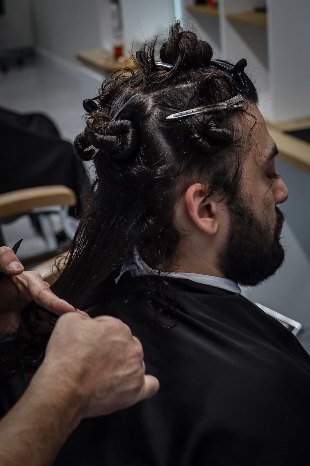 A stylist’s hands sectioning long curly hair with clips, the client in a black cape
