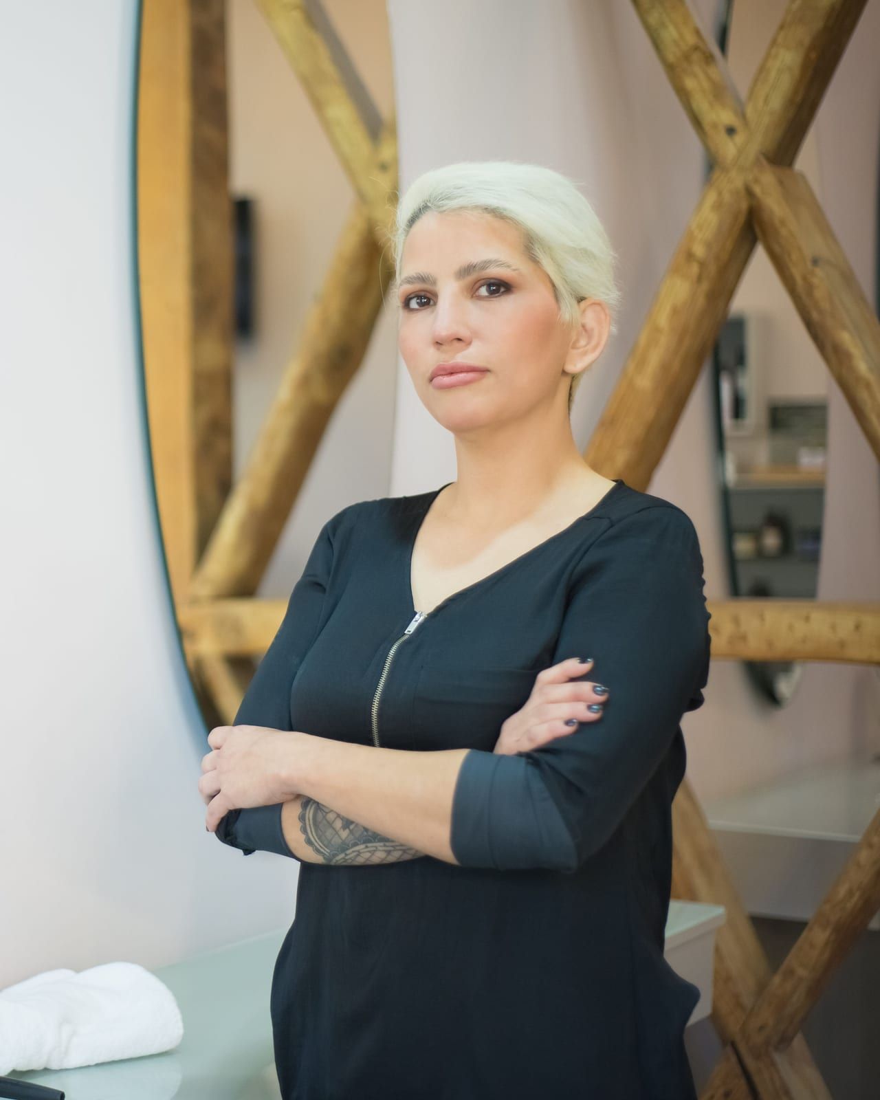 A hairdresser with short platinum hair, arms folded, beside a round salon mirror
