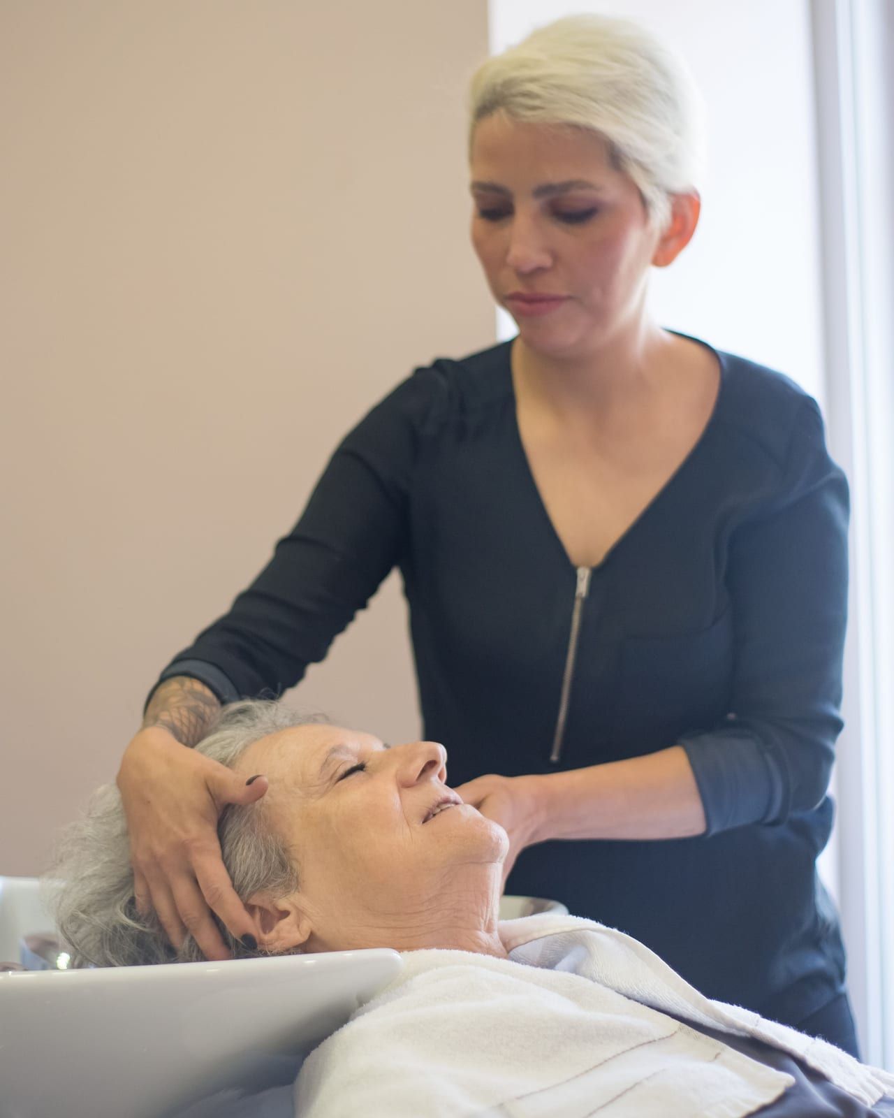 A hairdresser with short platinum hair massaging an older client’s scalp at a basin by a bright window