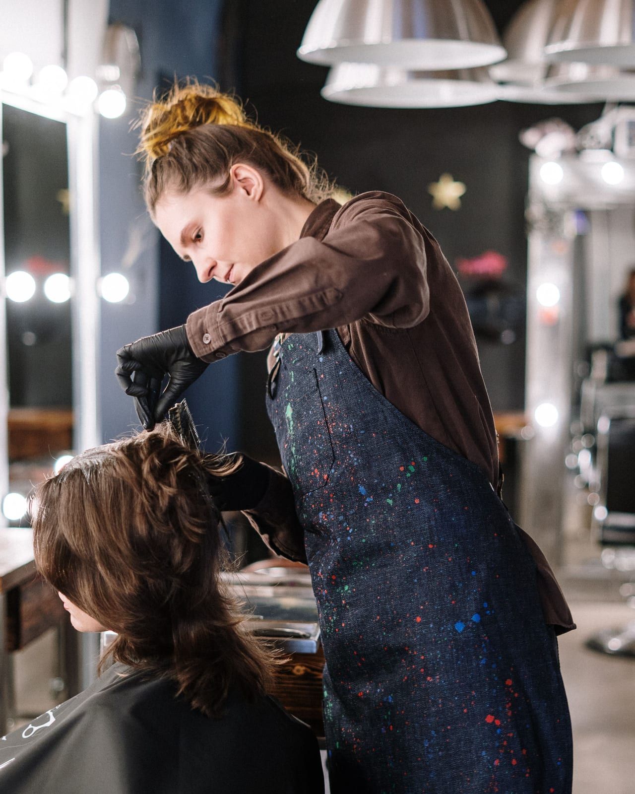 A hairdresser in a paint-flecked apron and black gloves brushing color through a client’s hair