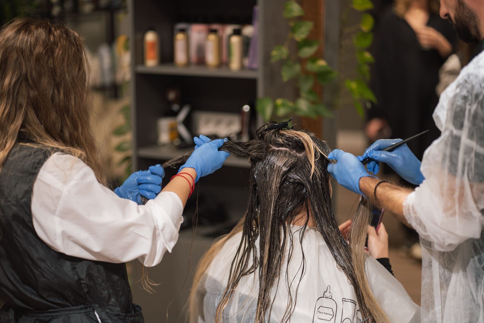 Two stylists in blue gloves painting color through a client’s long hair, shelves of products behind
