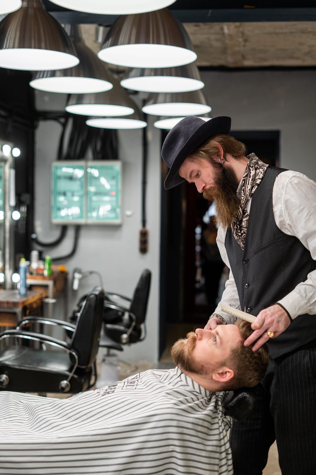 A barber in a hat and waistcoat trimming a reclining client’s beard