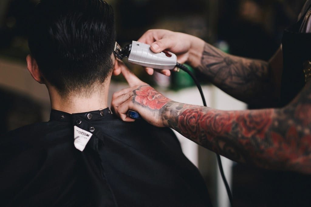 A barber with tattooed arms using clippers on a short cut